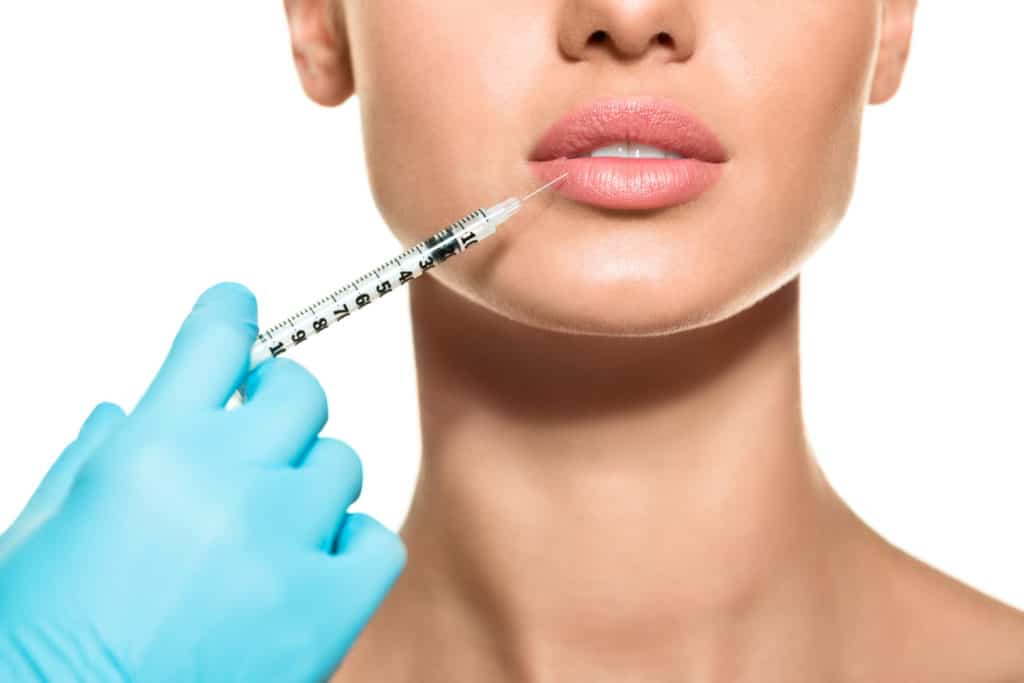 Close up of hand wearing surgical glowe holding a syringe injecting dermal filler in female lips.